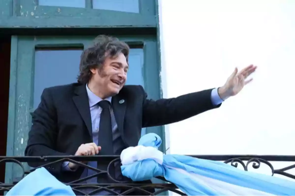 A smiling man in a dark suit waves from a balcony decorated with a light blue and white flag.