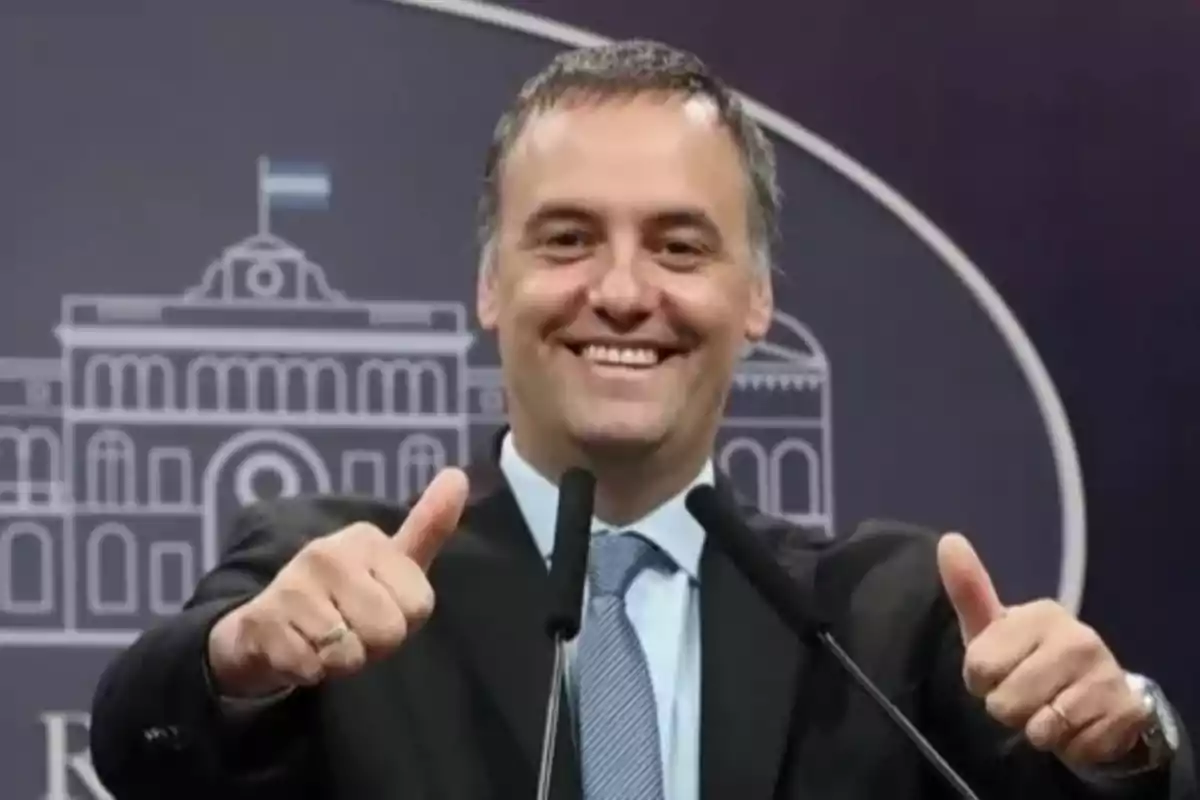 Un hombre sonriente con traje y corbata levanta ambos pulgares frente a un fondo con un dibujo de un edificio. Un hombre sonriente con traje y corbata levanta ambos pulgares frente a un fondo con un dibujo de un edificio.