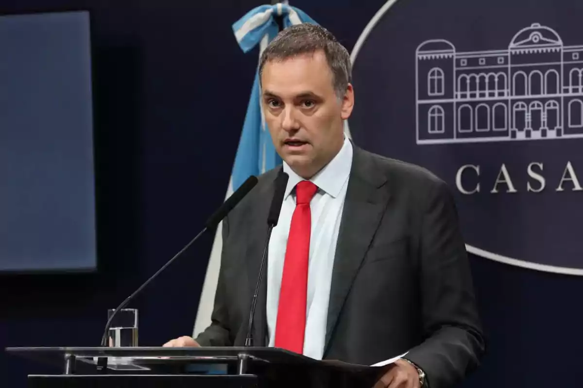 Un hombre en traje con corbata roja hablando en un podio con un micrófono y una bandera de fondo.