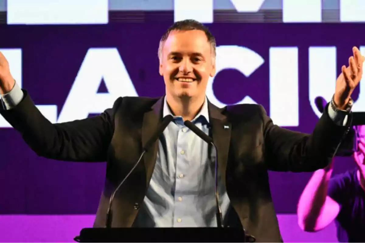 A smiling man in a suit and blue jersey raises his arms on a stage with a purple background and white letters. A smiling man in a suit and blue jersey raises his arms on a stage with a purple background and white letters.