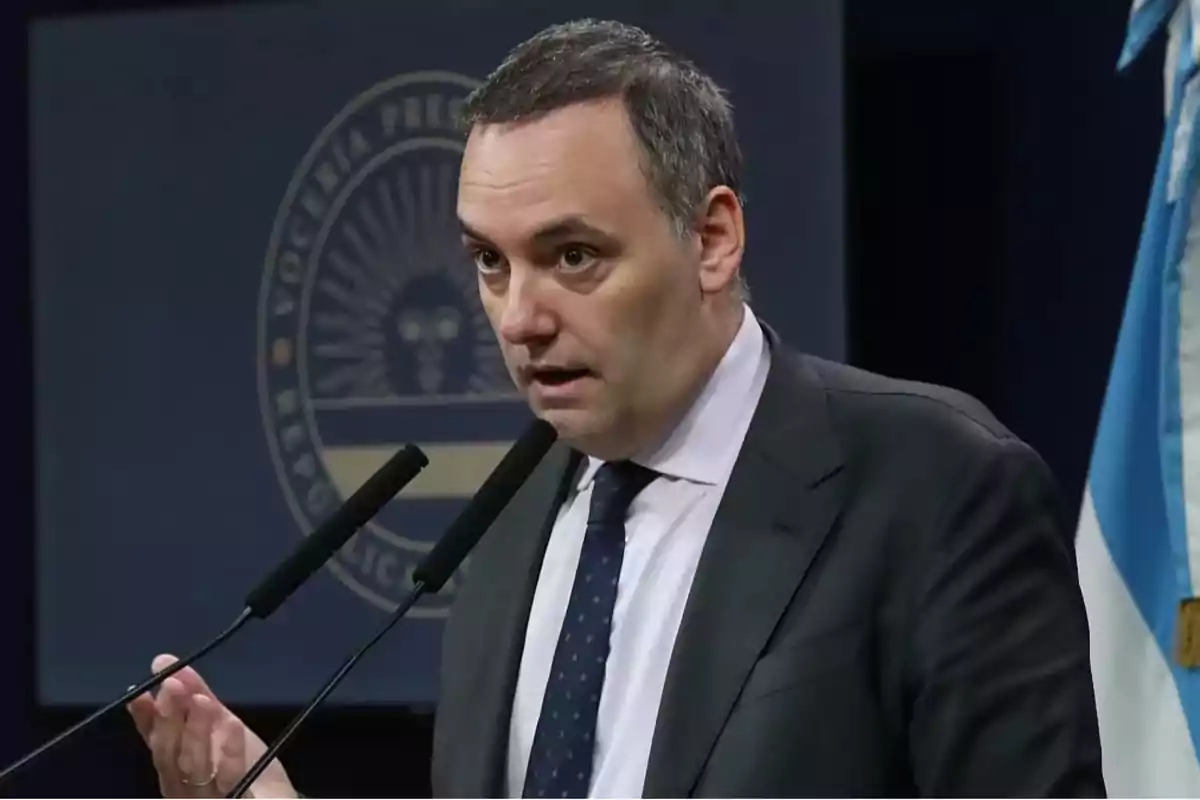 Un hombre en traje hablando en una conferencia de prensa con un micrófono frente a él y una bandera a su lado.