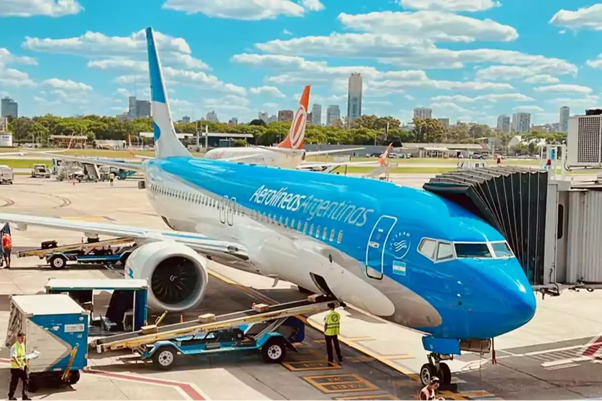 Un avión de Aerolíneas Argentinas estacionado en un aeropuerto con personal de tierra trabajando alrededor y un cielo azul con nubes en el fondo.