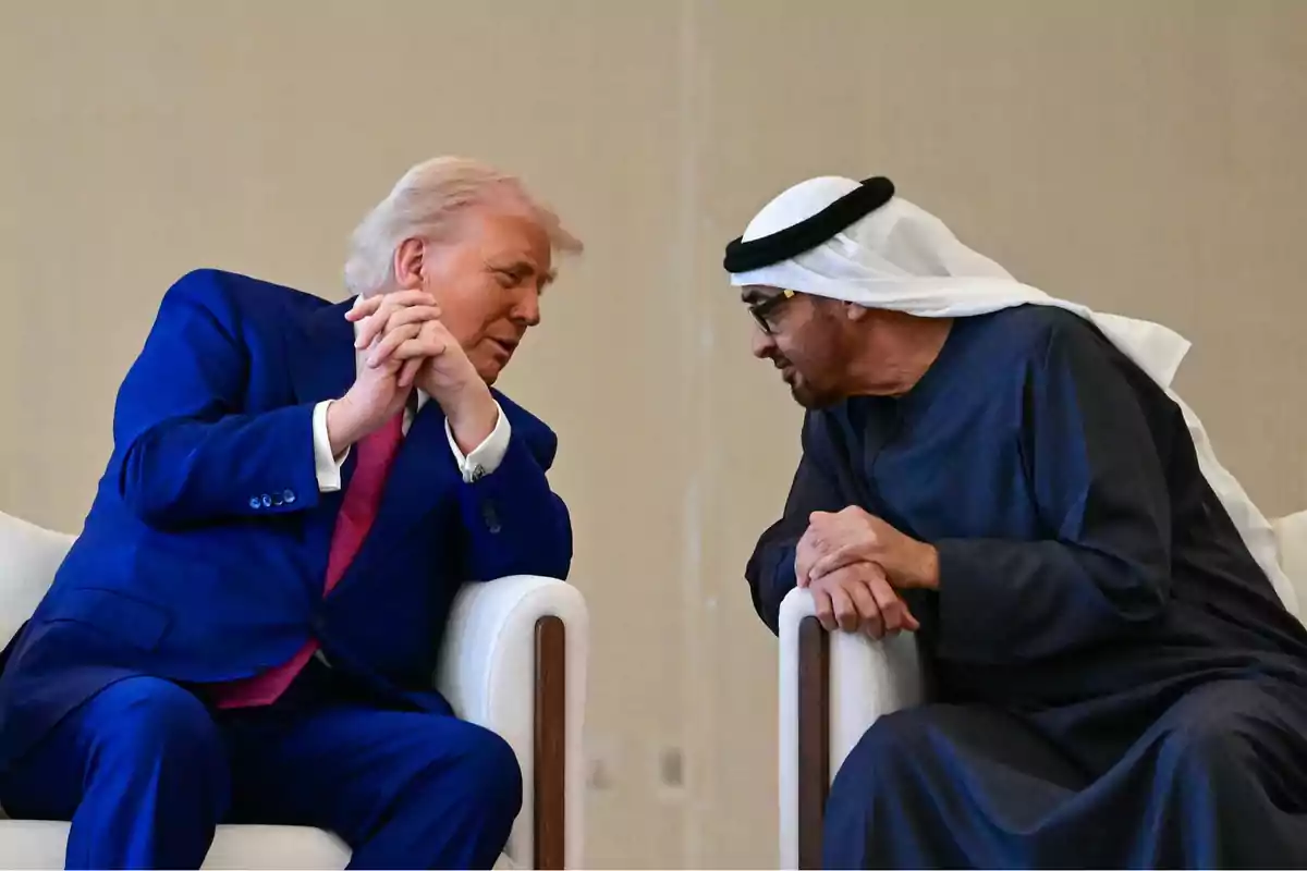Dos hombres sentados en sillones blancos conversan de cerca, uno lleva traje azul y corbata rosa, el otro viste túnica tradicional árabe y turbante blanco. Dos hombres sentados en sillones blancos conversan de cerca, uno lleva traje azul y corbata rosa, el otro viste túnica tradicional árabe y turbante blanco.
