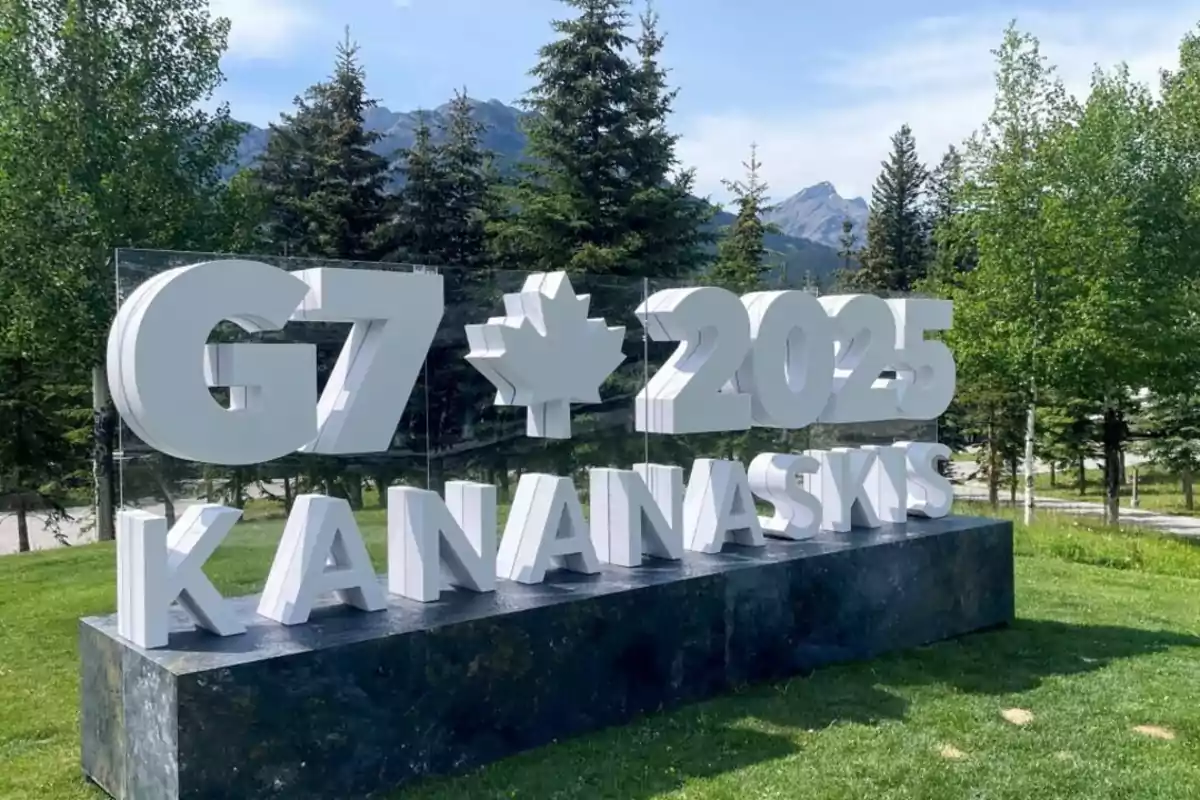 White letter structure that reads G7 2025 Kananaskis with a maple leaf symbol in a natural setting with trees and mountains in the background White letter structure that reads G7 2025 Kananaskis with a maple leaf symbol in a natural setting with trees and mountains in the background