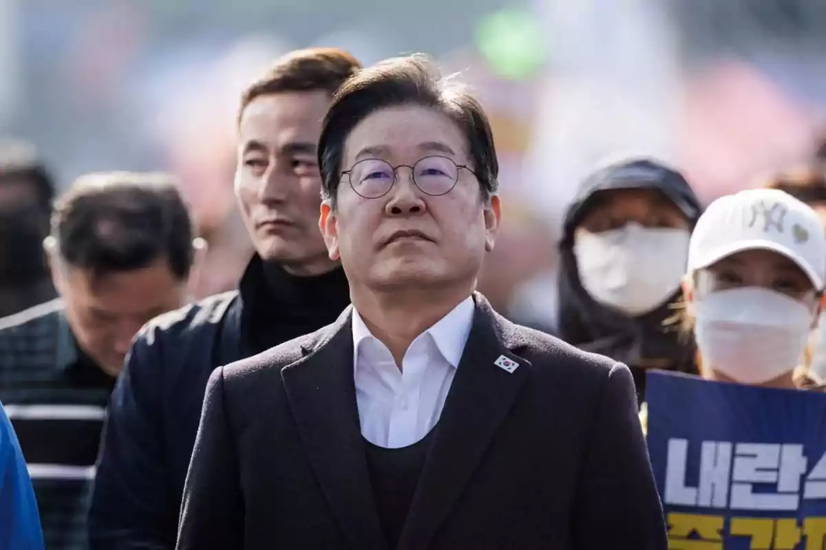 A group of people at an outdoor event, with a man in front wearing glasses and a dark suit, while others behind are wearing masks. A group of people at an outdoor event, with a man in front wearing glasses and a dark suit, while others behind are wearing masks.