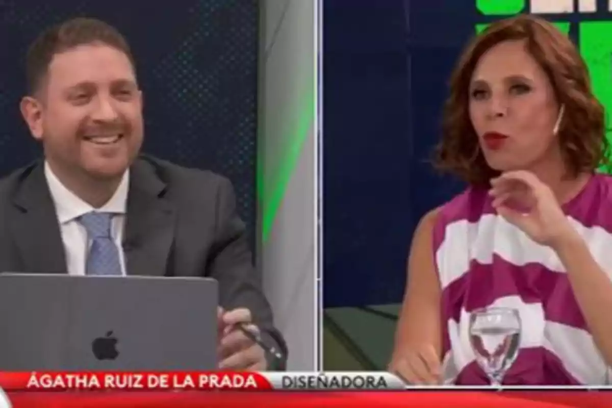 Dos personas conversando en un programa de televisión, una de ellas con un traje y corbata y la otra con un vestido a rayas, con un rótulo en la parte inferior que dice 