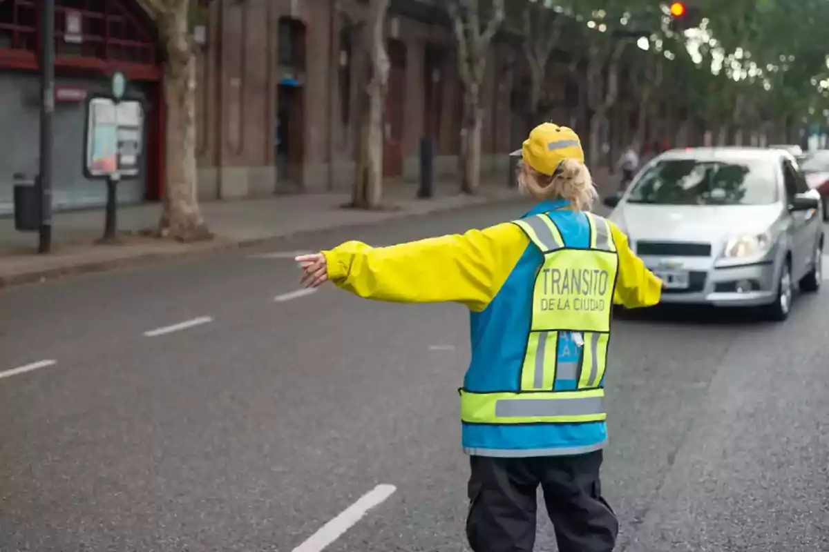 Una agente de tránsito con chaleco amarillo y azul dirige el tráfico en una calle urbana mientras un automóvil se aproxima.