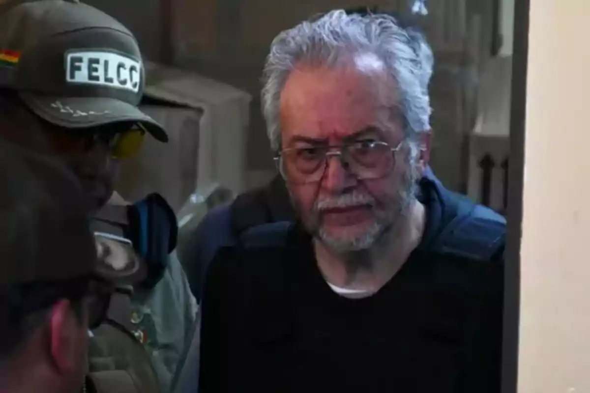 Un hombre de cabello canoso y barba, con gafas, es escoltado por un oficial de policía con uniforme y gorra que tiene la insignia Un hombre de cabello canoso y barba, con gafas, es escoltado por un oficial de policía con uniforme y gorra que tiene la insignia