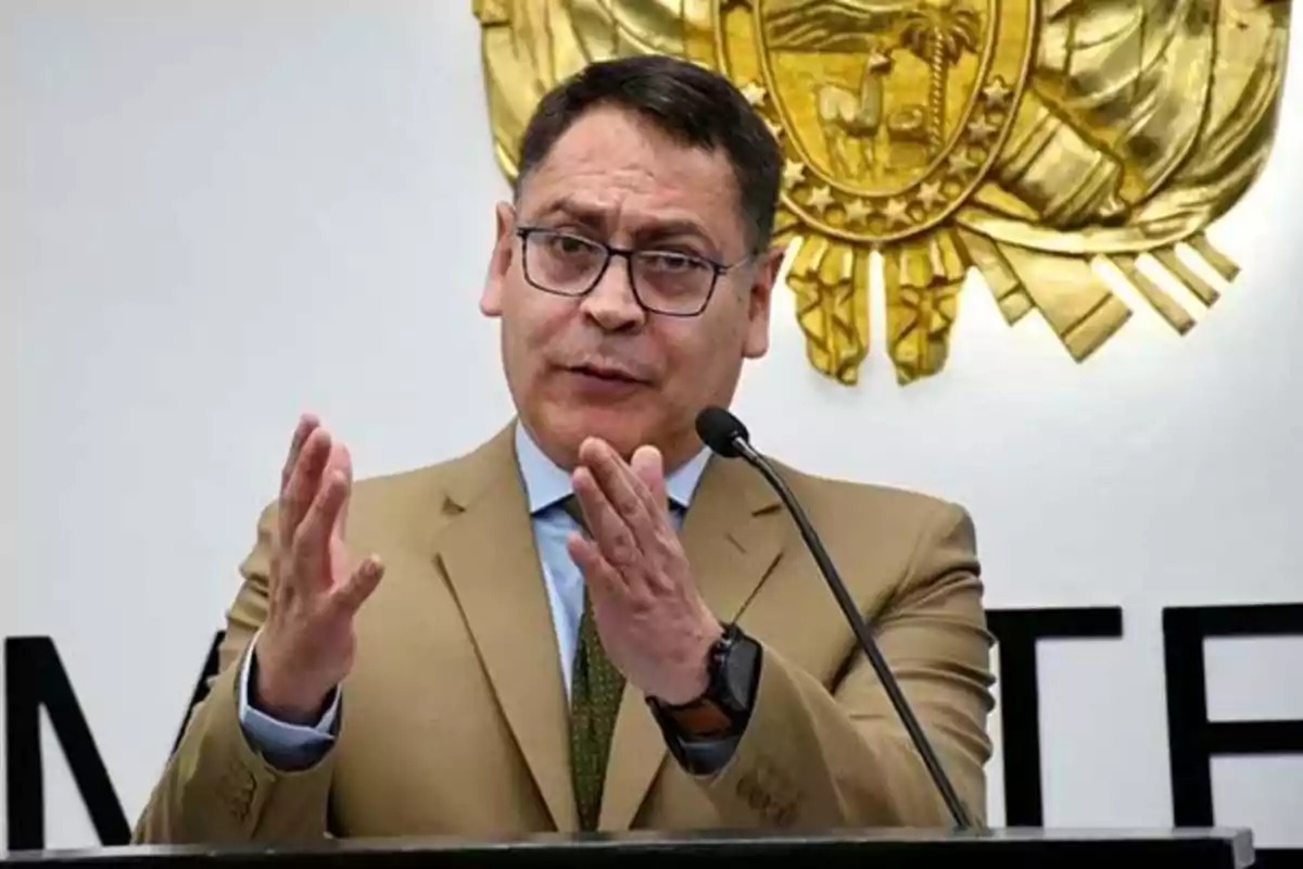 Un hombre con gafas y traje marrón habla en un micrófono frente a un escudo dorado en la pared.