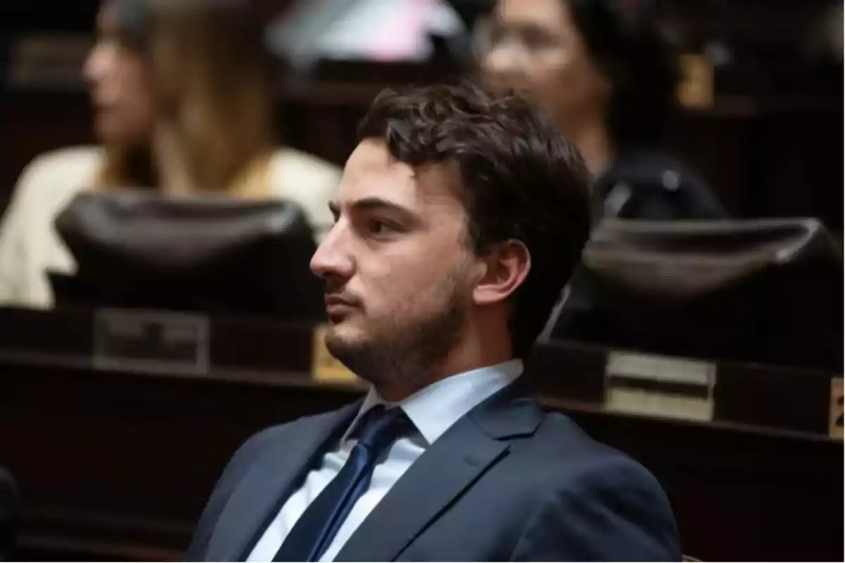 Un hombre con traje y corbata está sentado en un auditorio o sala de conferencias.