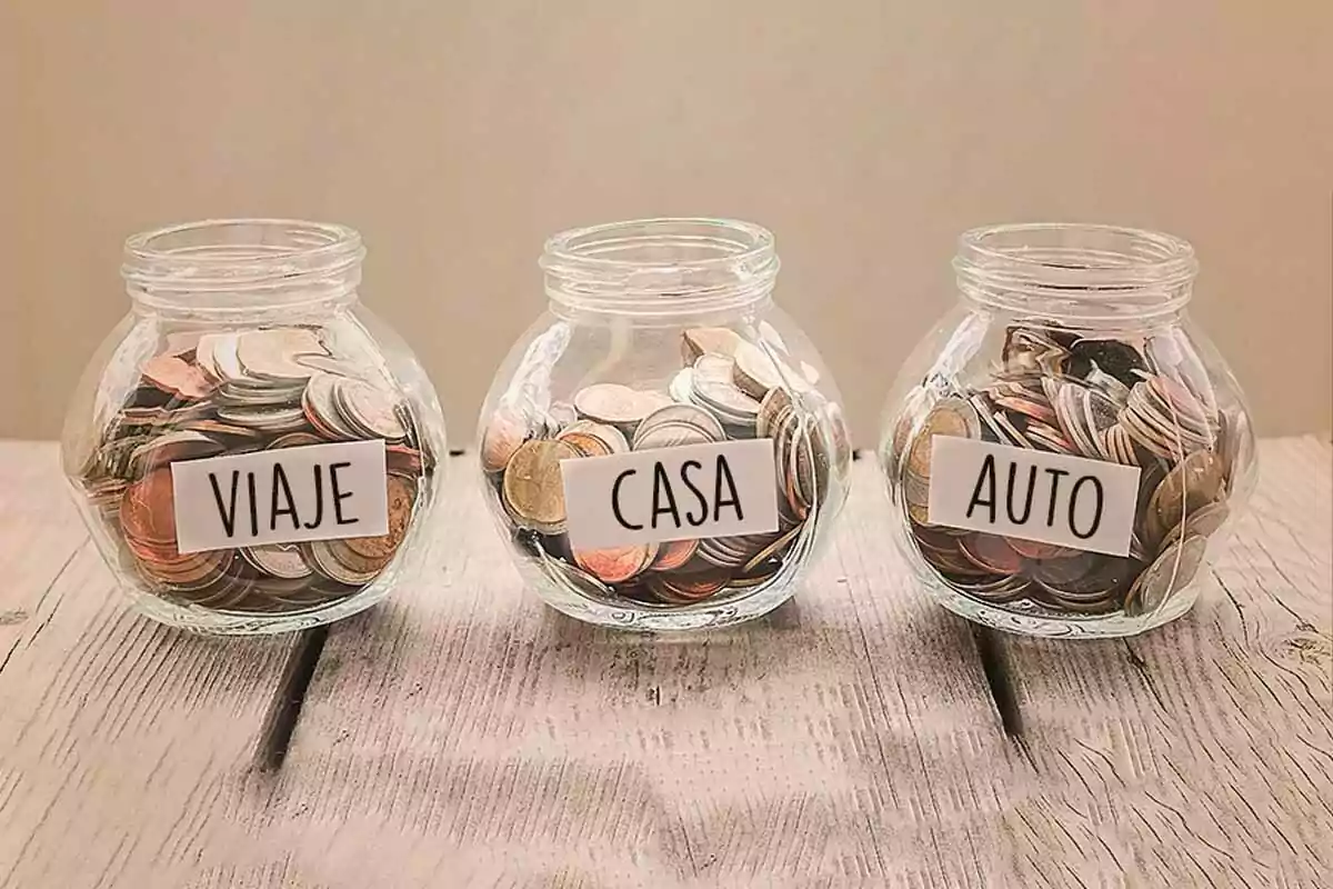 Tres frascos de vidrio llenos de monedas con etiquetas que dicen viaje casa y auto sobre una mesa de madera clara Tres frascos de vidrio llenos de monedas con etiquetas que dicen viaje casa y auto sobre una mesa de madera clara