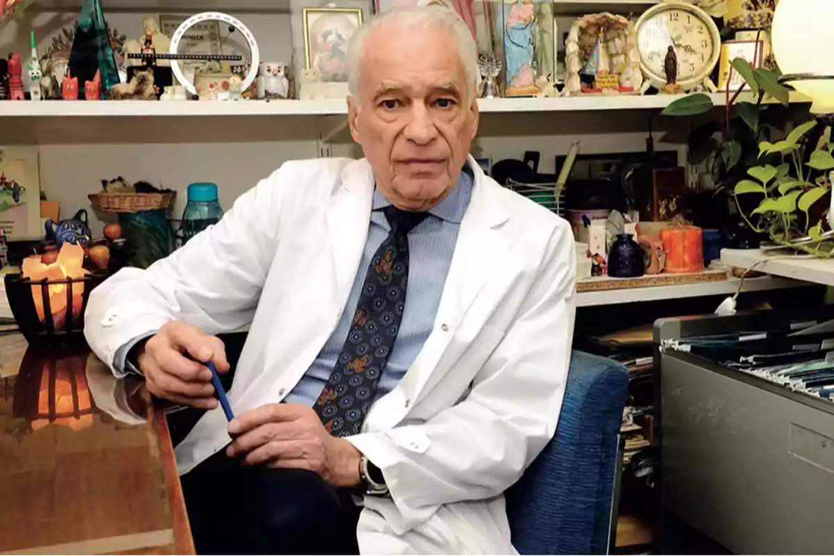 An elderly man in a white robe is sitting in an office filled with decorative objects and plants.