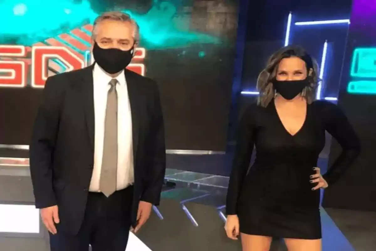 Dos personas con mascarillas posan en un set de televisión iluminado con luces de neón y un fondo con el logotipo de un programa.