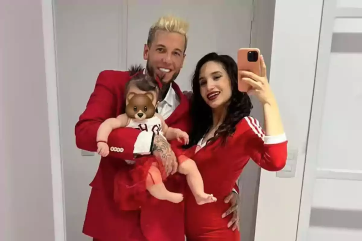 A smiling couple dressed in red poses for a mirror selfie while holding a baby with a bear emoji covering its face.
