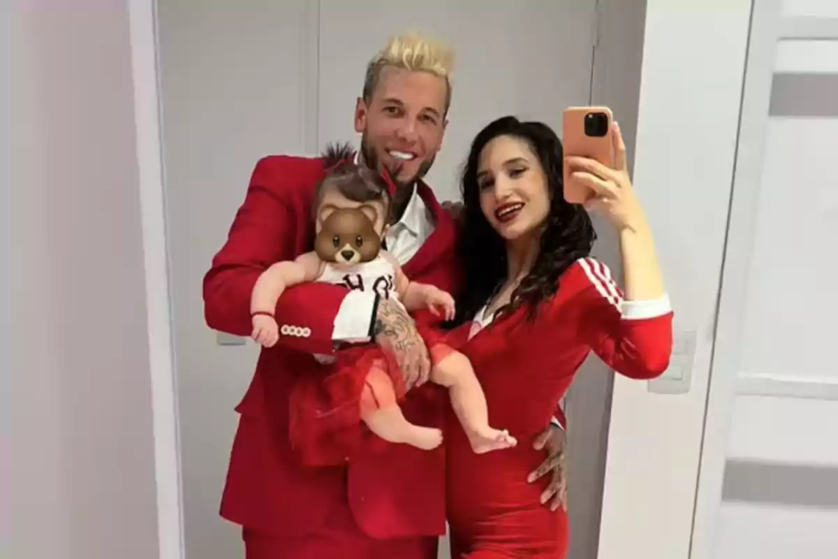 Una pareja vestida de rojo posa para una selfie en el espejo mientras sostienen a un bebé con un emoji de oso en la cara.