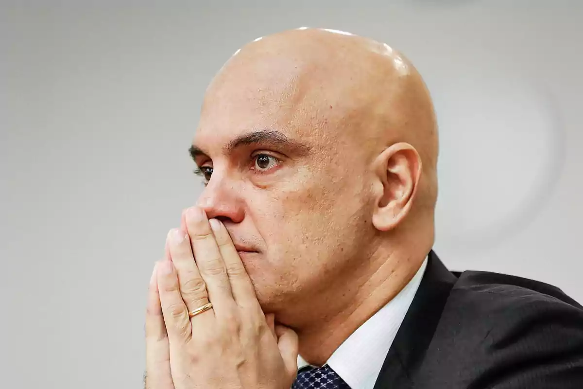 A bald man in a suit and tie is thoughtful with his hands together in front of his face.