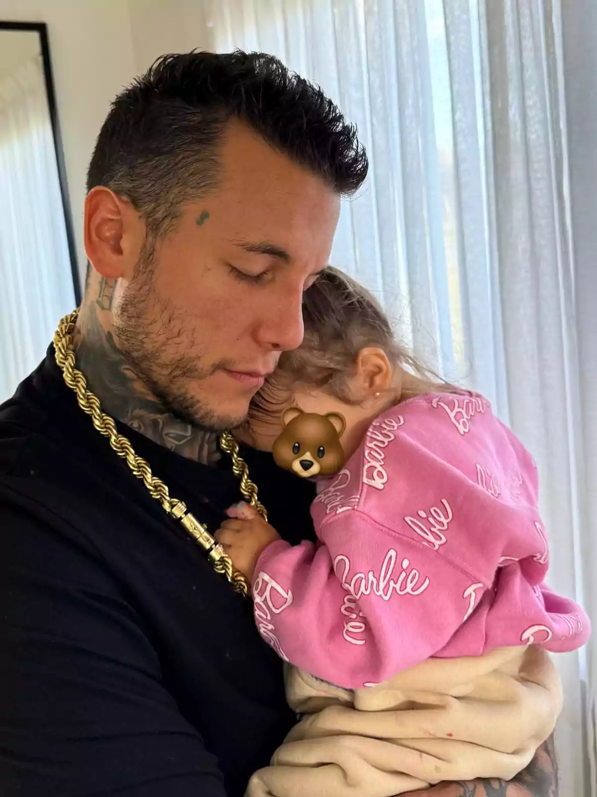 A man with tattoos and a gold chain lovingly holds a small child in a pink sweater. A man with tattoos and a gold chain lovingly holds a small child in a pink sweater.