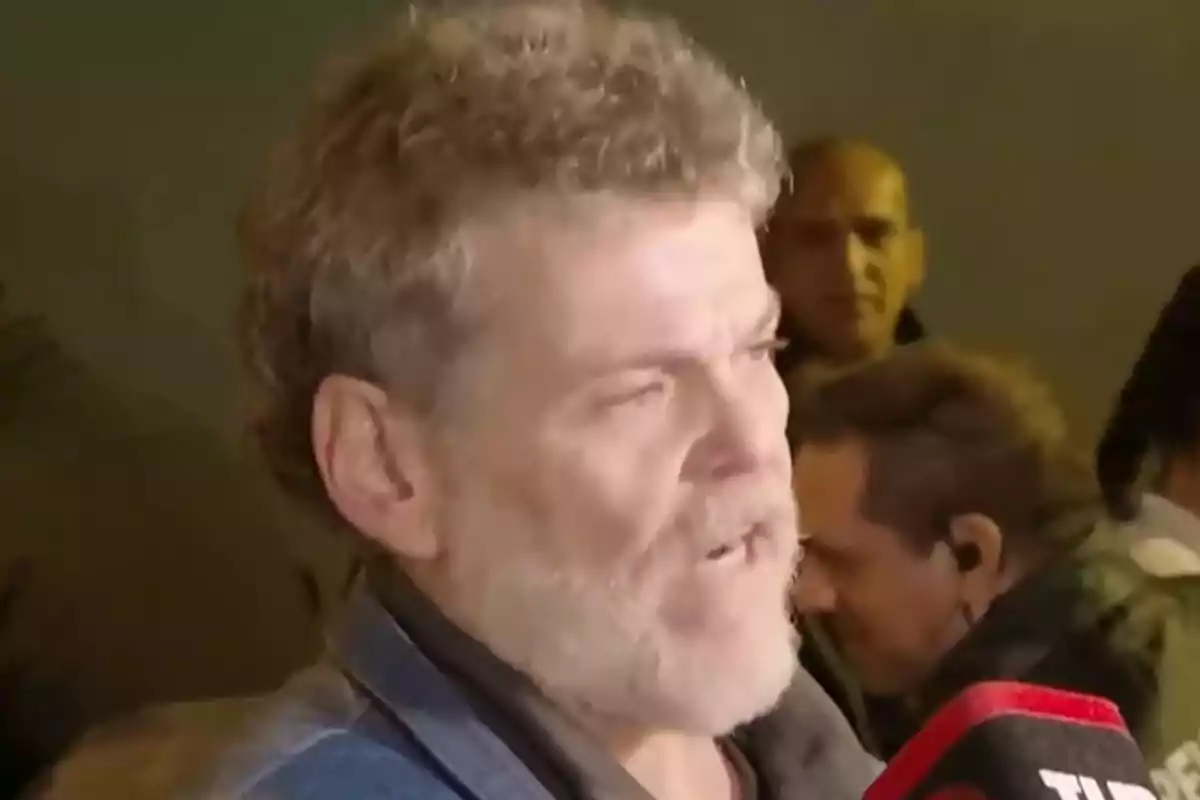 A man with a beard and curly hair speaks into a microphone surrounded by people in an outdoor setting. A man with a beard and curly hair speaks into a microphone surrounded by people in an outdoor setting.