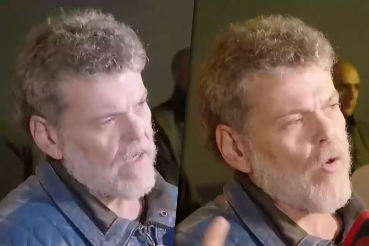 A man with a beard and curly hair is speaking indoors, wearing a blue jacket.