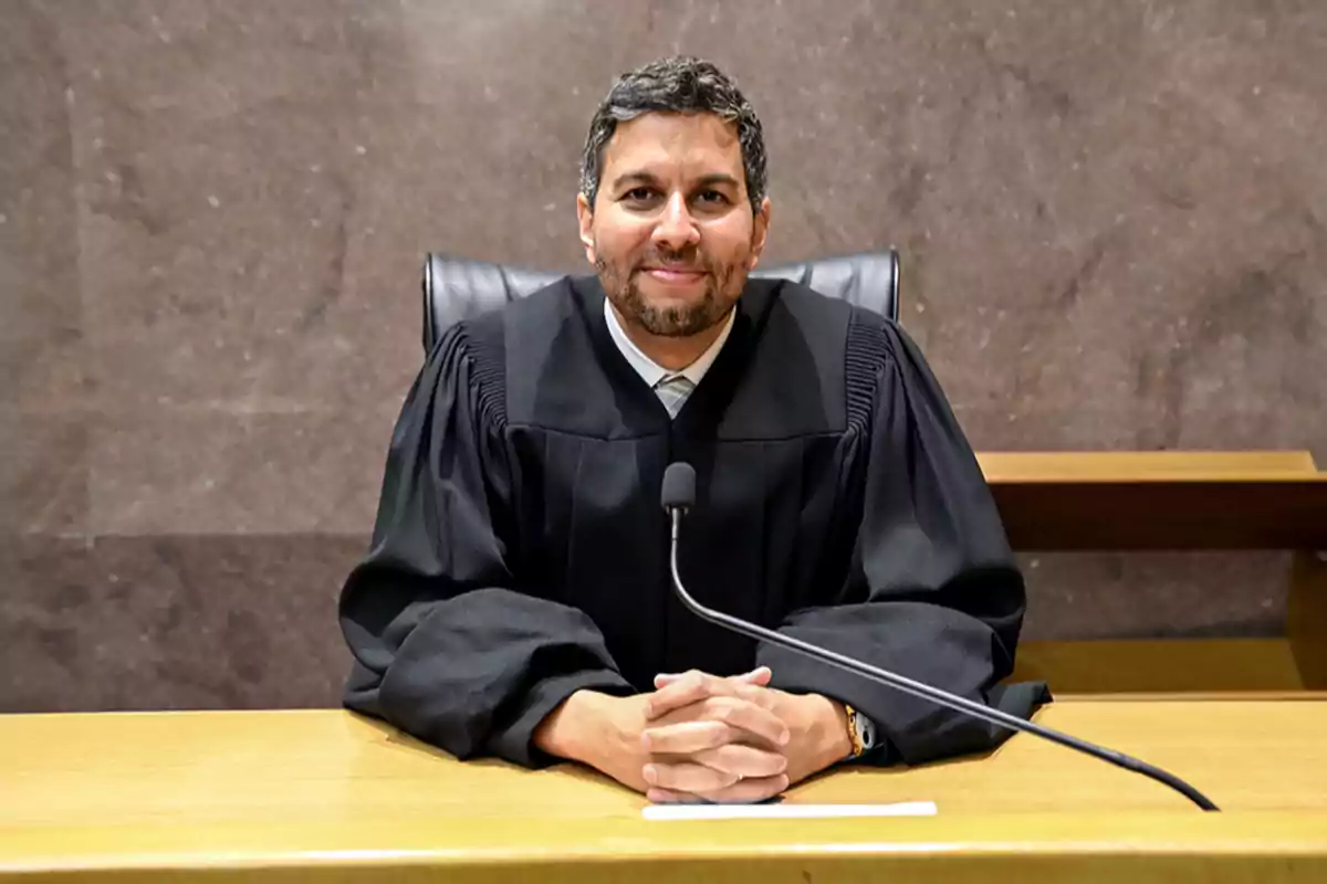 A judge sitting at his desk with hands clasped and wearing a black robe.