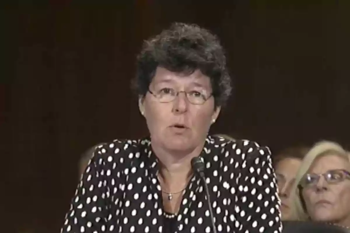 A person with glasses and curly hair speaks into a microphone, wearing a blouse with white polka dots on a dark background. A person with glasses and curly hair speaks into a microphone, wearing a blouse with white polka dots on a dark background.