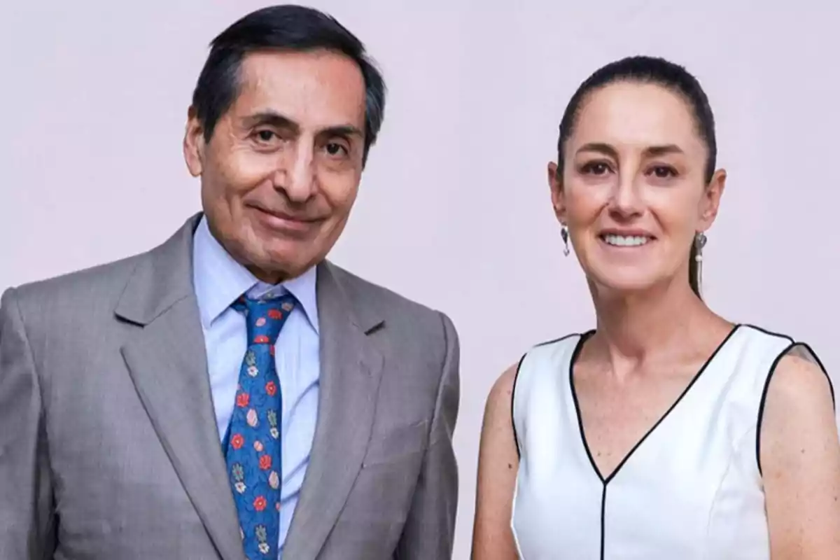 Dos personas posando juntas, una vestida con traje gris y corbata azul, y la otra con blusa blanca sin mangas.