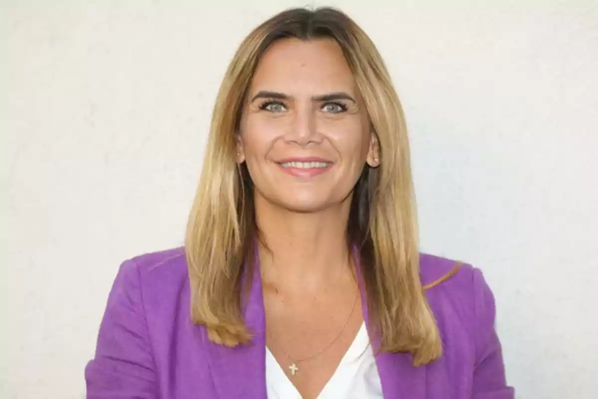 Blonde-haired woman with straight hair wearing a purple jacket and white blouse, smiling in front of a light-colored wall