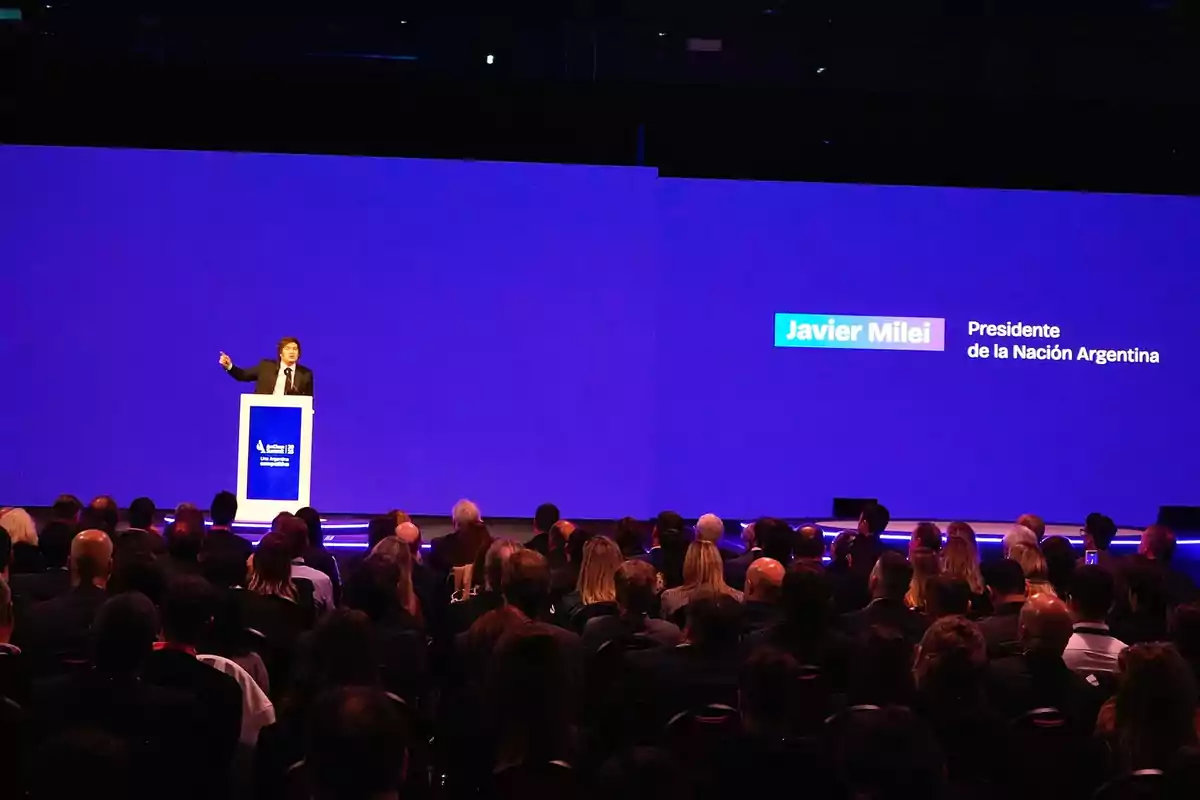 Una persona hablando en un escenario frente a una audiencia con una pantalla azul de fondo. Una persona hablando en un escenario frente a una audiencia con una pantalla azul de fondo.