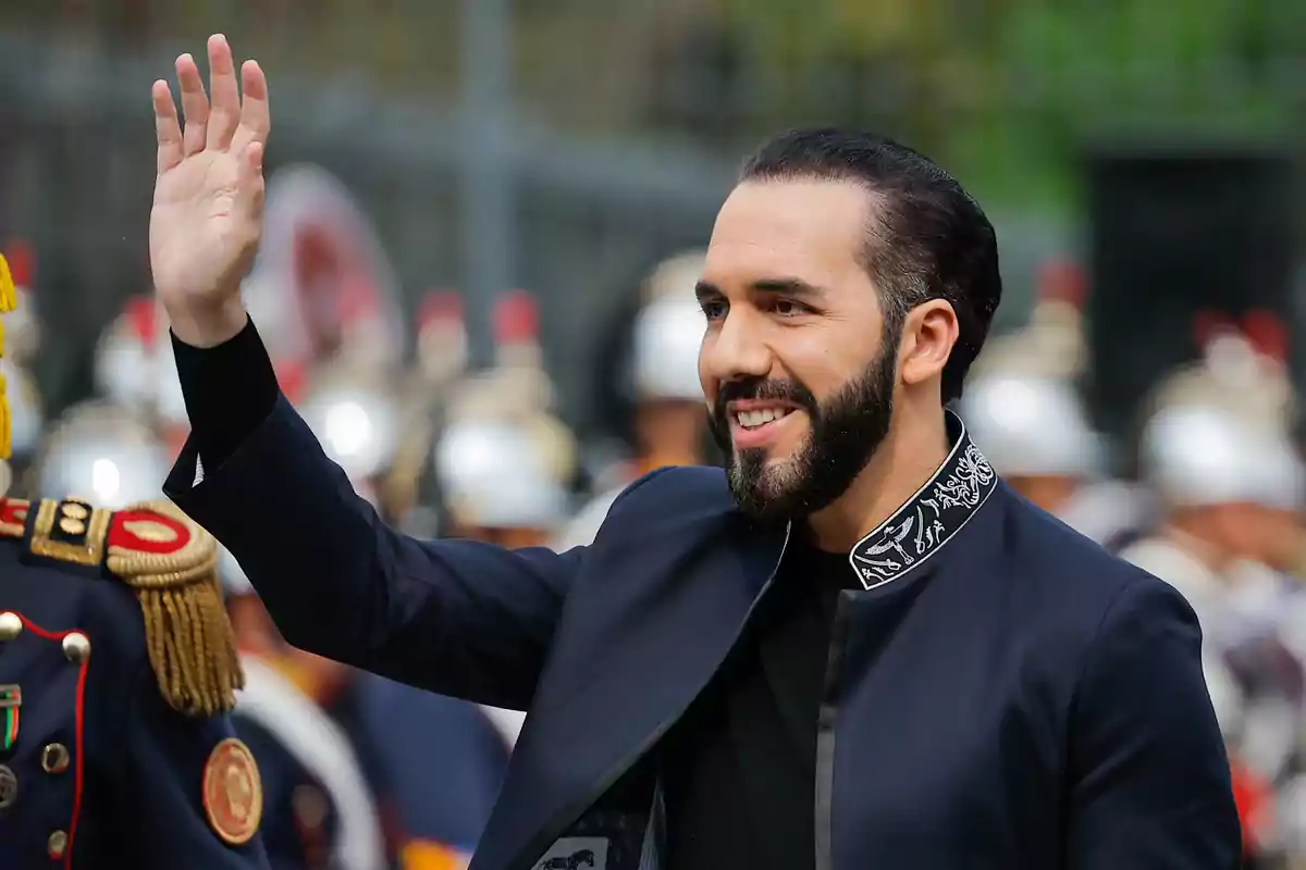 Un hombre con barba y cabello oscuro, vestido con una chaqueta formal, saluda con la mano levantada mientras sonríe, con un fondo de personas uniformadas.