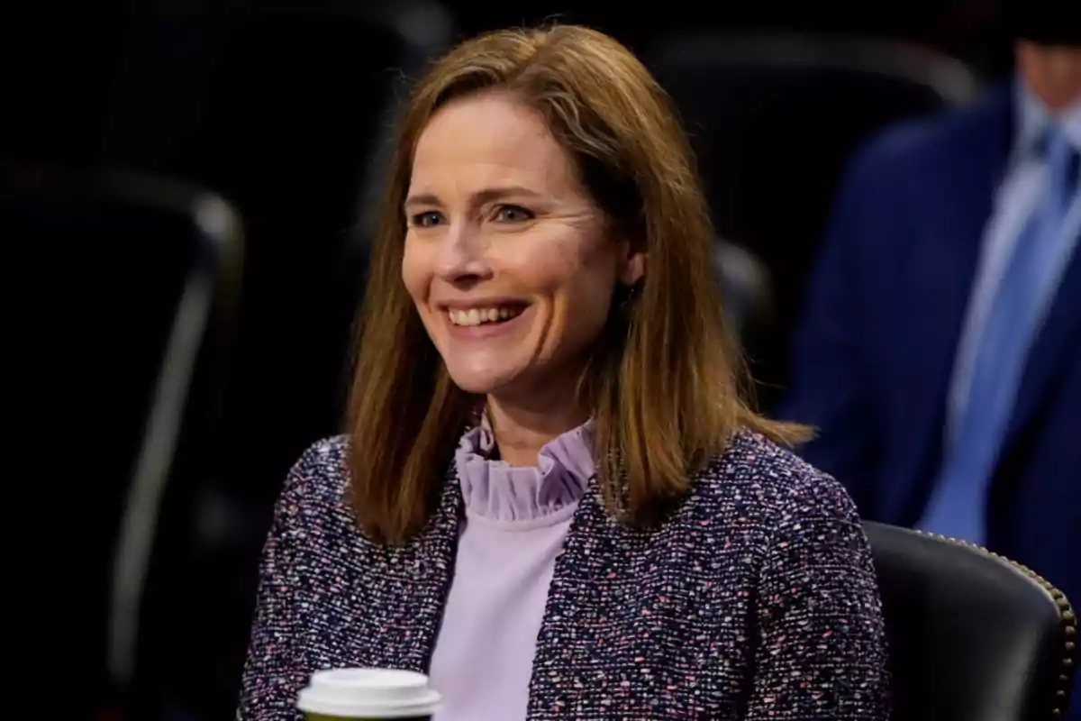 A smiling woman with brown hair and a tweed jacket holds a cup of coffee in a formal setting. A smiling woman with brown hair and a tweed jacket holds a cup of coffee in a formal setting.
