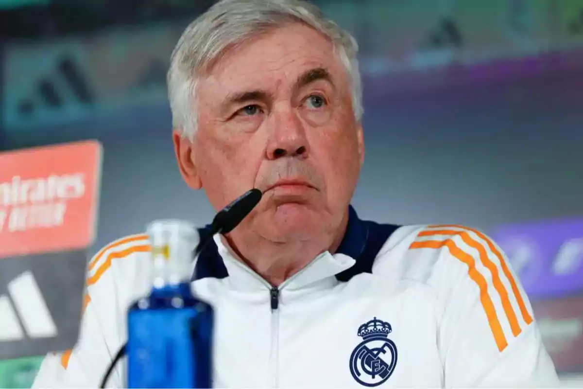 A man in a white sports jacket with orange details, sitting in front of a microphone at a press conference.