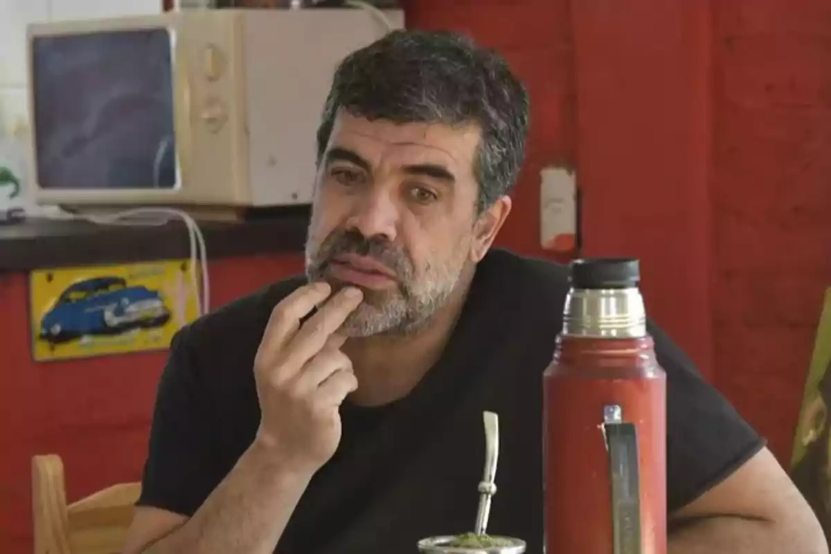 Dark-haired, bearded man sitting at a table with a thermos and a mate, in a homey setting with red walls and a microwave in the background. Dark-haired, bearded man sitting at a table with a thermos and a mate, in a homey setting with red walls and a microwave in the background.