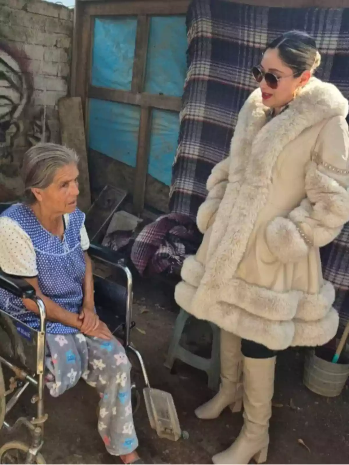 Una mujer mayor en silla de ruedas conversa con otra mujer que lleva un abrigo de piel y gafas de sol en un entorno al aire libre.