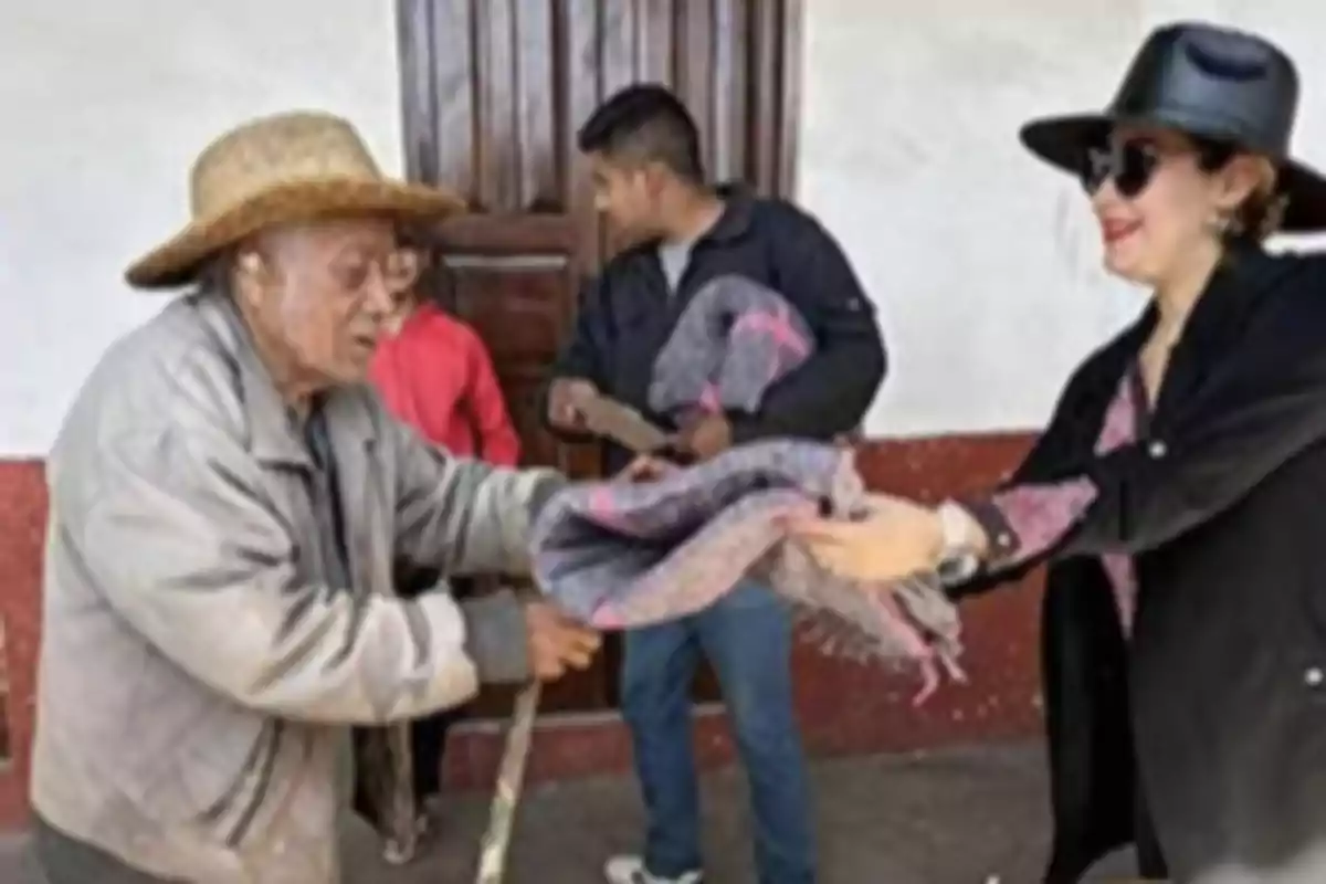 Una persona mayor recibe una cobija de una mujer mientras otras personas observan en el fondo.