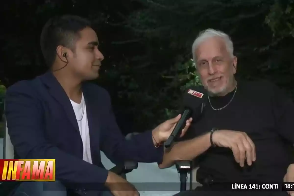 Un entrevistador sostiene un micrófono mientras conversa con un hombre mayor de cabello blanco en un entorno al aire libre. Un entrevistador sostiene un micrófono mientras conversa con un hombre mayor de cabello blanco en un entorno al aire libre.