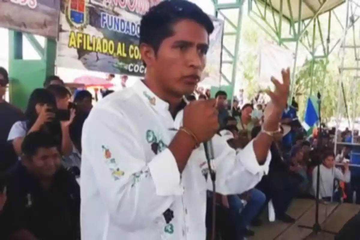 Un hombre joven con camisa blanca decorada habla en un micrófono frente a una audiencia en un evento al aire libre.