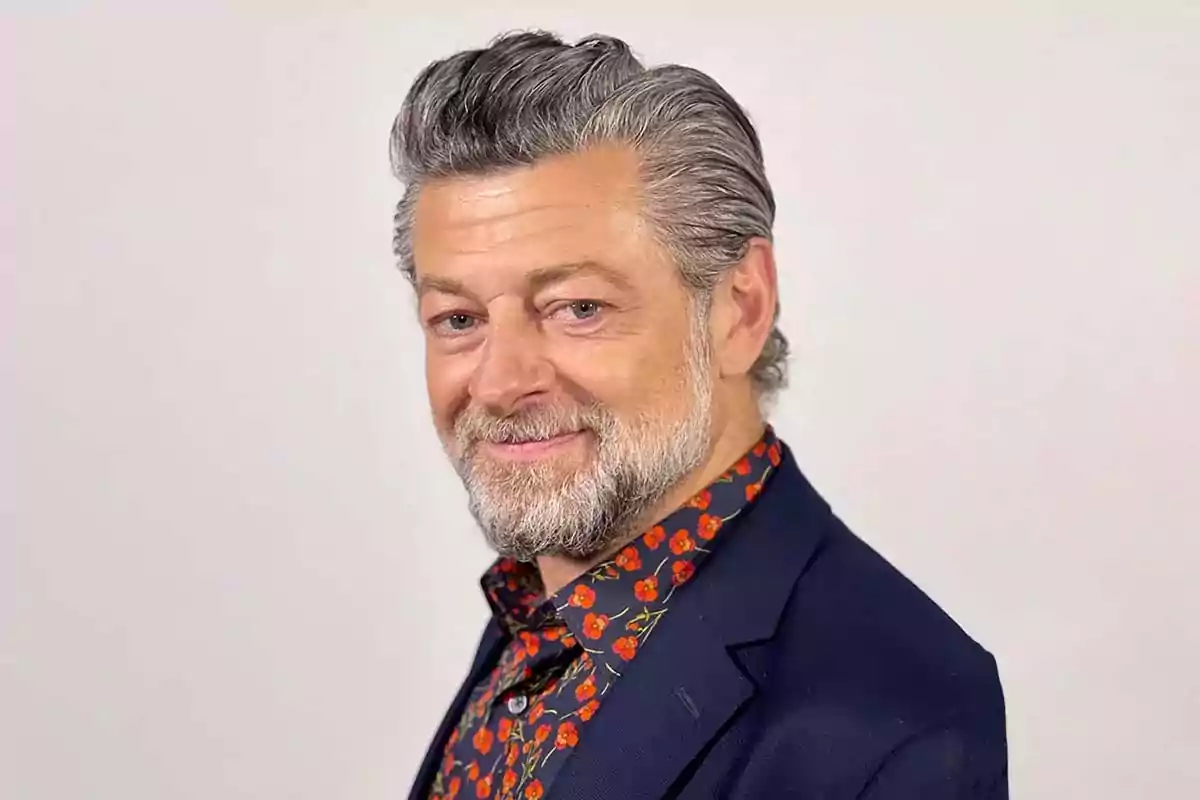 Gray-haired, bearded man wearing a floral jersey and blue blazer smiling in front of a light background Gray-haired, bearded man wearing a floral jersey and blue blazer smiling in front of a light background