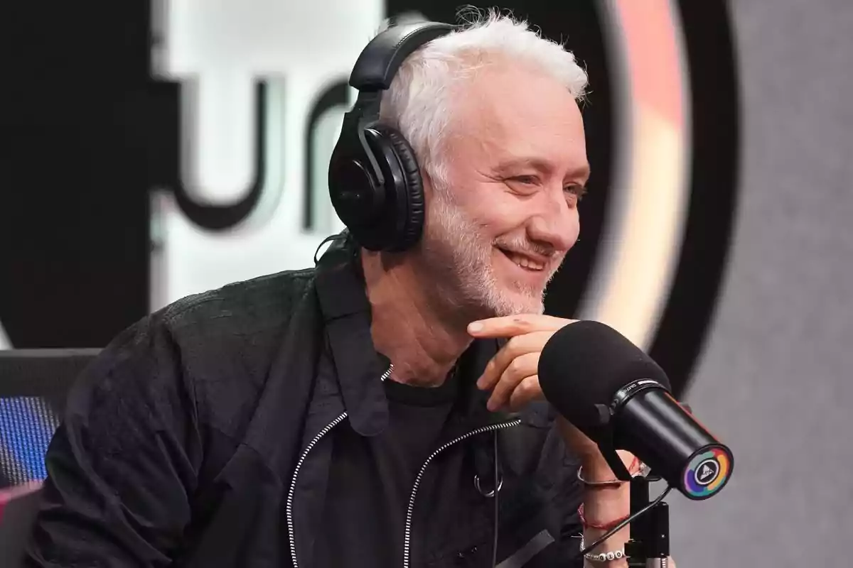 Gray-haired man with a short beard wearing headphones and smiling while speaking in front of a microphone in a radio studio