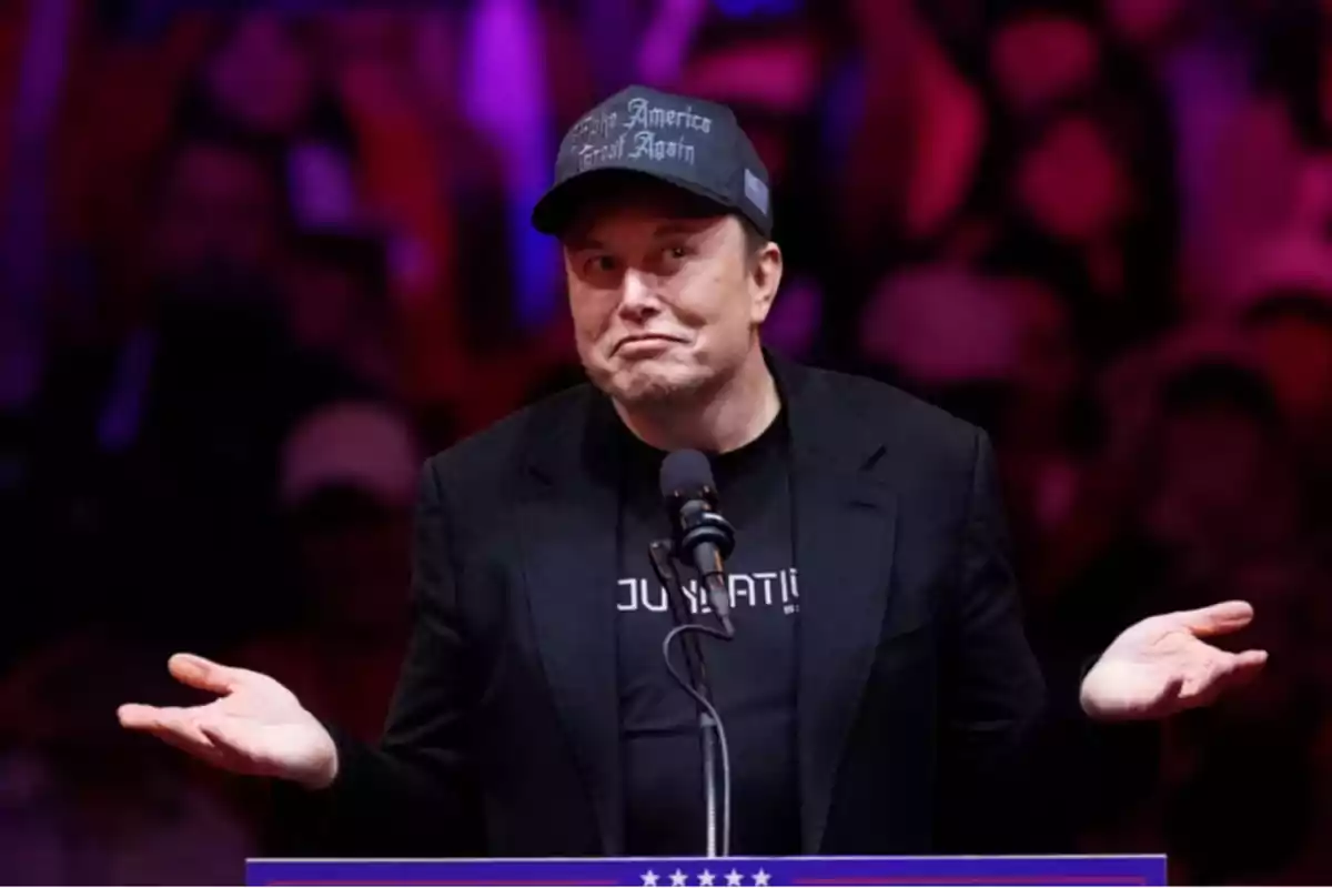 Un hombre con gorra y chaqueta negra gesticula con las manos levantadas frente a un micrófono en un escenario iluminado en tonos rojos y morados.