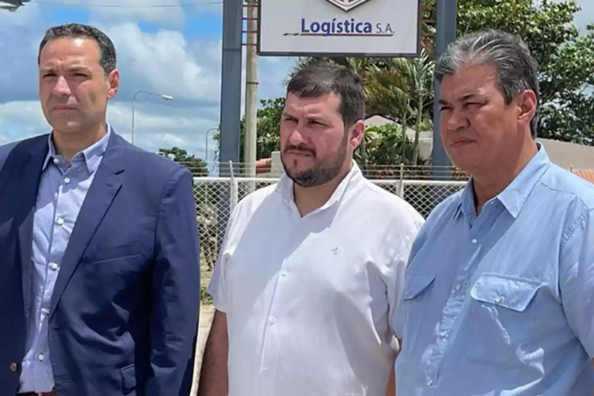 Tres hombres de pie al aire libre frente a un cartel que dice "Logística S.A." con cielo parcialmente nublado de fondo.