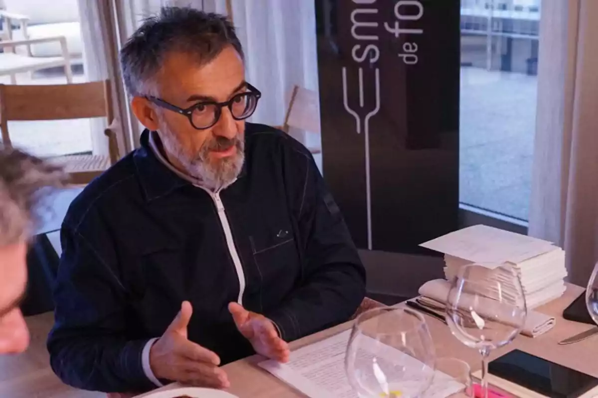 A man with glasses and a beard is sitting at a table, talking and gesturing with his hands. There are glasses and papers on the table.
