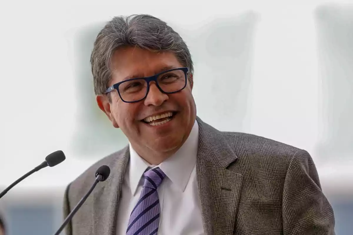A man with glasses and a tie smiles while speaking into a microphone. A man with glasses and a tie smiles while speaking into a microphone.