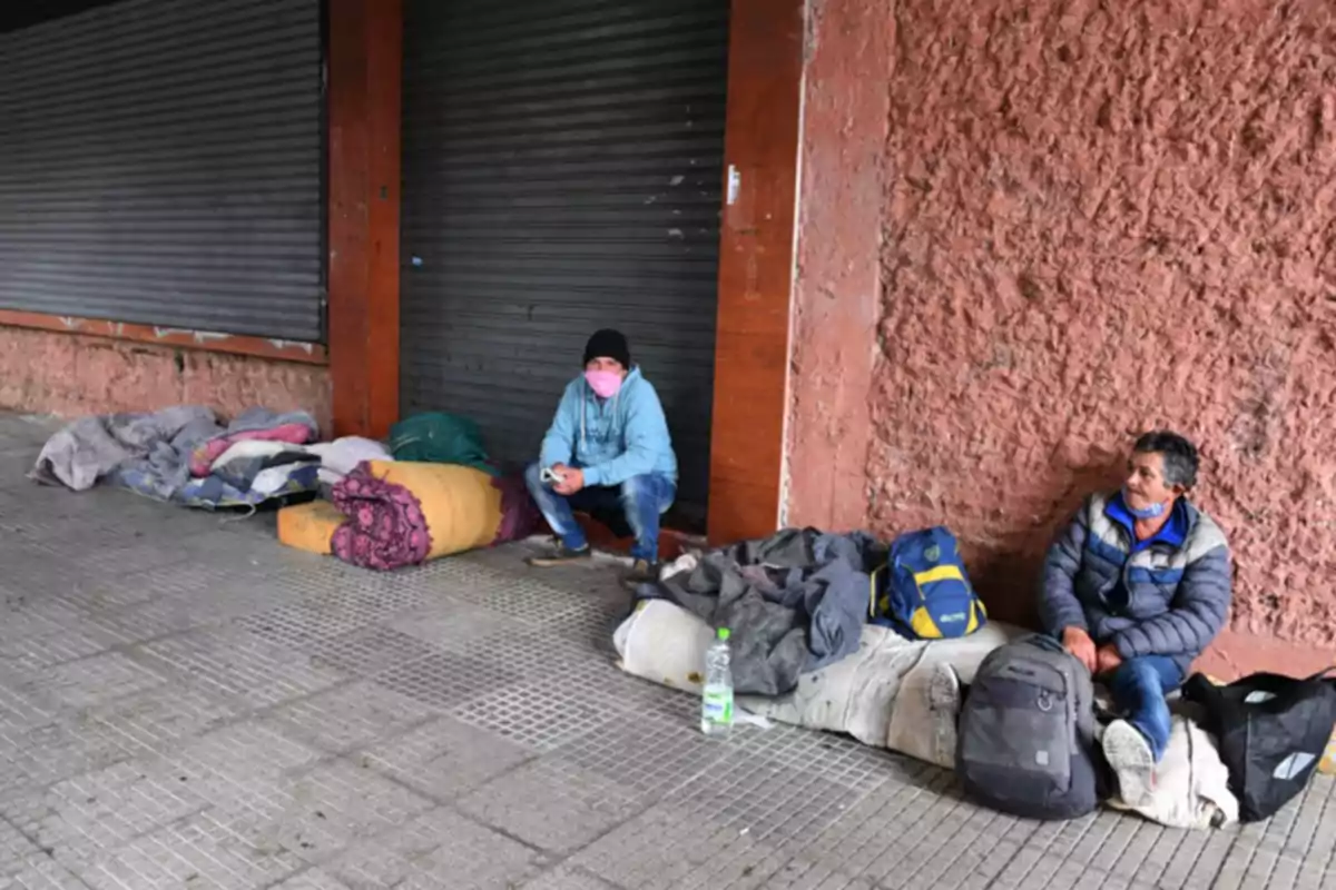 Two people sitting on the sidewalk next to a brick wall, surrounded by backpacks and blankets. Two people sitting on the sidewalk next to a brick wall, surrounded by backpacks and blankets.
