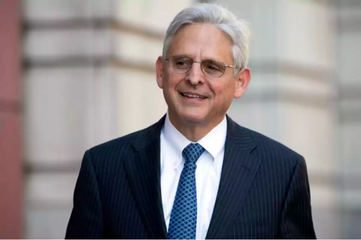 Gray-haired man with glasses and a dark suit smiles while standing in front of a building. Gray-haired man with glasses and a dark suit smiles while standing in front of a building.