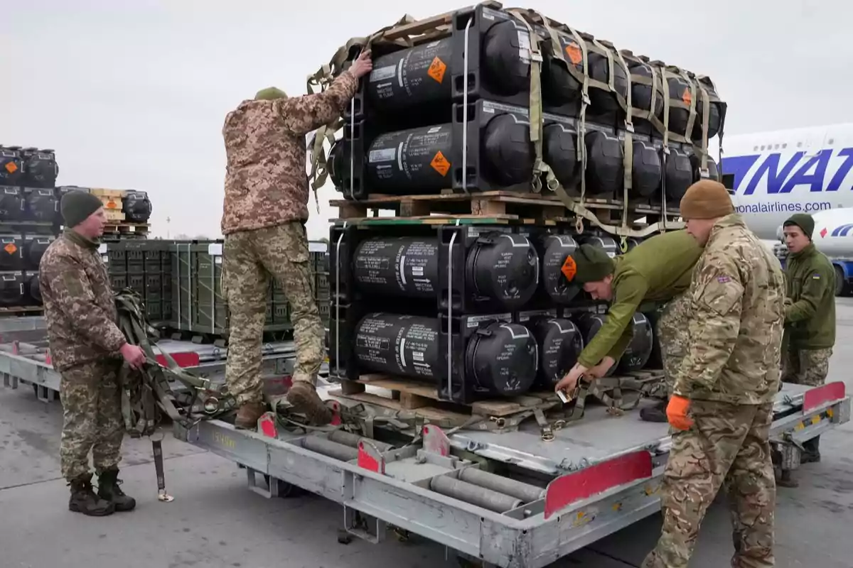 Soldados en uniforme militar cargan y aseguran contenedores de equipo en una plataforma de transporte en un aeropuerto.