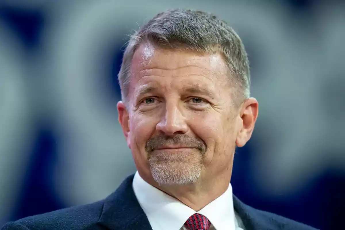 Hombre de mediana edad con cabello corto y barba, vestido con traje y corbata, sonriendo frente a un fondo desenfocado.