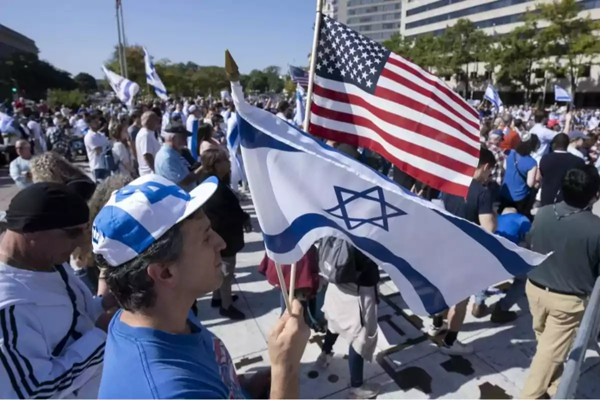 Hombre sostiene banderas de Israel y Estados Unidos en una manifestación con muchas personas reunidas al aire libre Hombre sostiene banderas de Israel y Estados Unidos en una manifestación con muchas personas reunidas al aire libre