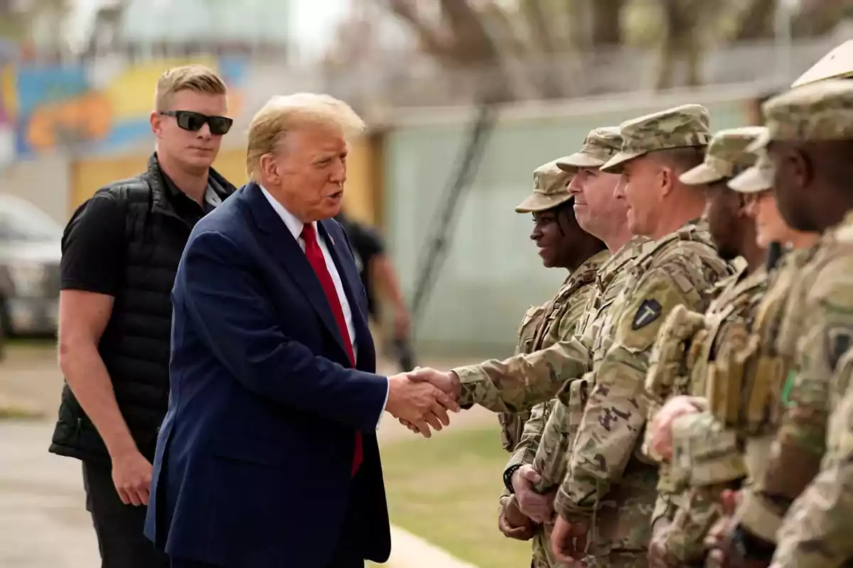 Un hombre de traje estrechando la mano a un grupo de militares en uniforme de camuflaje, mientras otra persona observa desde atrás. Un hombre de traje estrechando la mano a un grupo de militares en uniforme de camuflaje, mientras otra persona observa desde atrás.