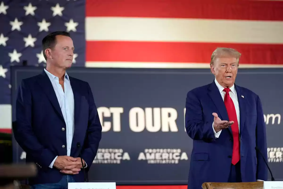 Dos hombres de pie en un escenario con una bandera de Estados Unidos de fondo, uno de ellos gesticulando mientras el otro observa.