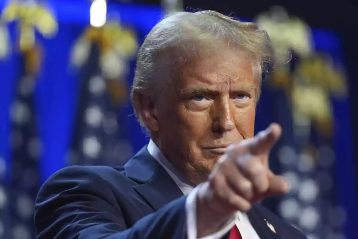 A man in a dark suit points forward with a serious expression, against a background of blurred flags. A man in a dark suit points forward with a serious expression, against a background of blurred flags.